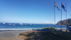 Uma praia com barcos na água, bandeiras e um afloramento rochoso.