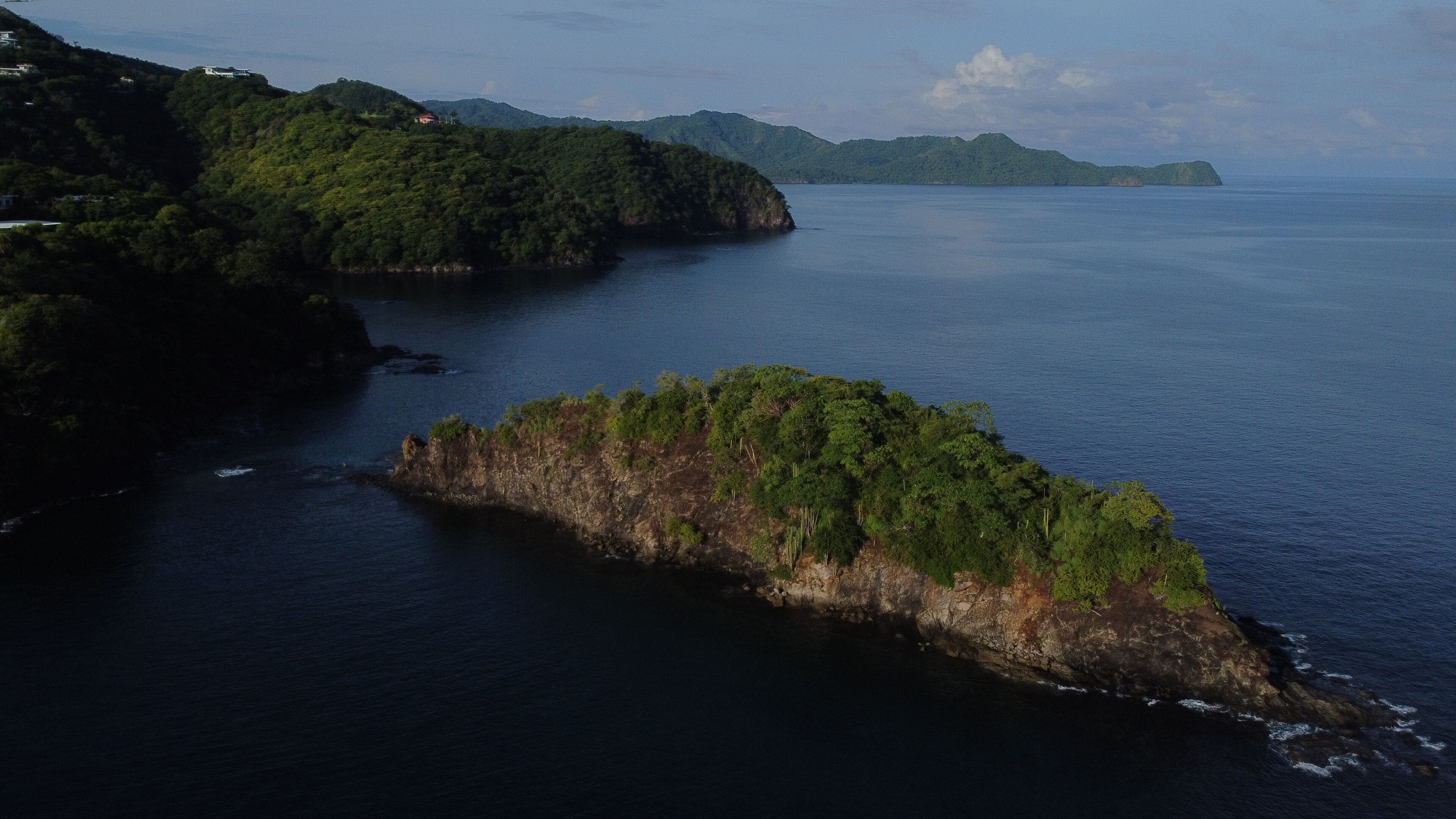 A coastal landscape with a prominent rocky island surrounded by water, with distant hills and a clear sky.