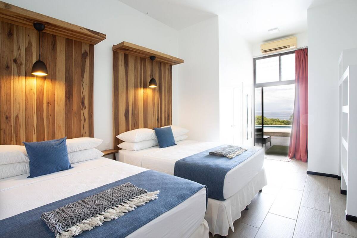 Habitació d'hotel amb dos llits, ambres de fusta i vista al mar a través d'una finestra gran.