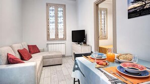 Ein möbliertes Wohnzimmer mit einer Couch, einem Esstisch gedeckt mit Tellern, Gläsern und einer Kanne Orangensaft.