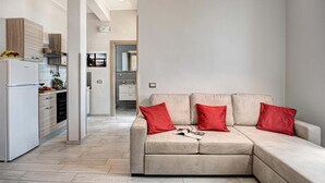Ein modernes Wohnzimmer mit einem beigefarbenen Sofa, roten Kissen und einer Küchenzeile mit Kühlschrank und Holzschränken.