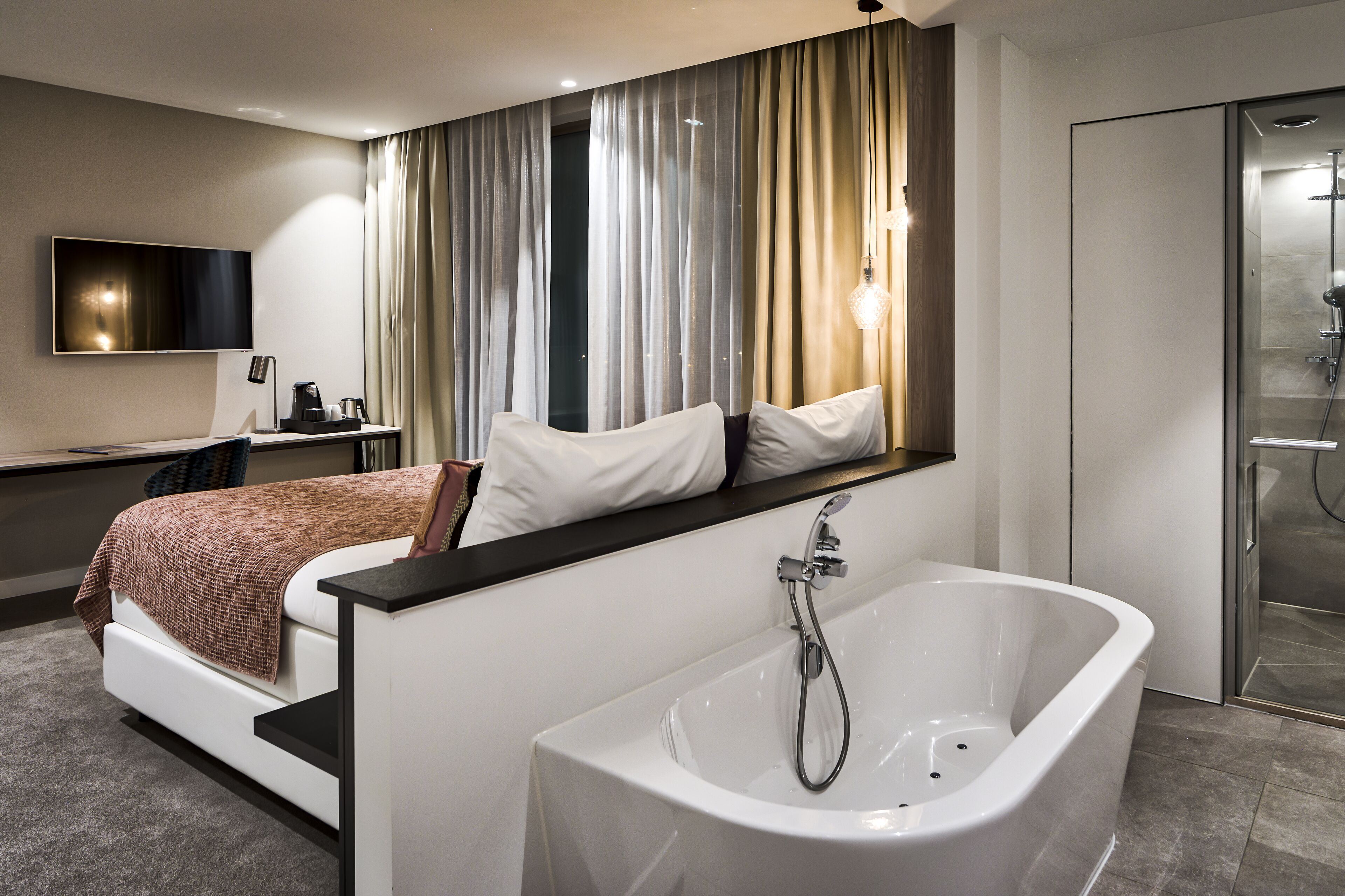A modern hotel room with a large bed, a built-in TV, a desk with a coffee maker, and a separate shower area.