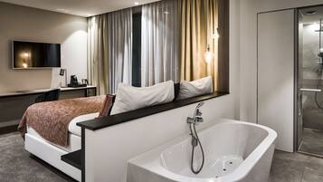 A modern hotel room with a large bed, a built-in TV, a desk with a coffee maker, and a separate shower area.