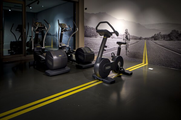 Un gimnasio con bicicletas estáticas y cintas de correr, un ventanal con vistas a una carretera y montañas, y un espejo que refleja el equipo.