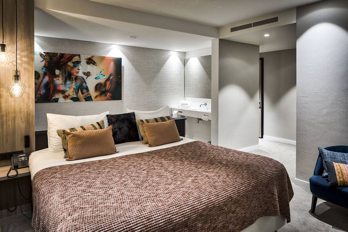 A modern bedroom with a large bed, a bedside table, a nightstand, a wall-mounted TV, and a bathroom with a sink and mirror.