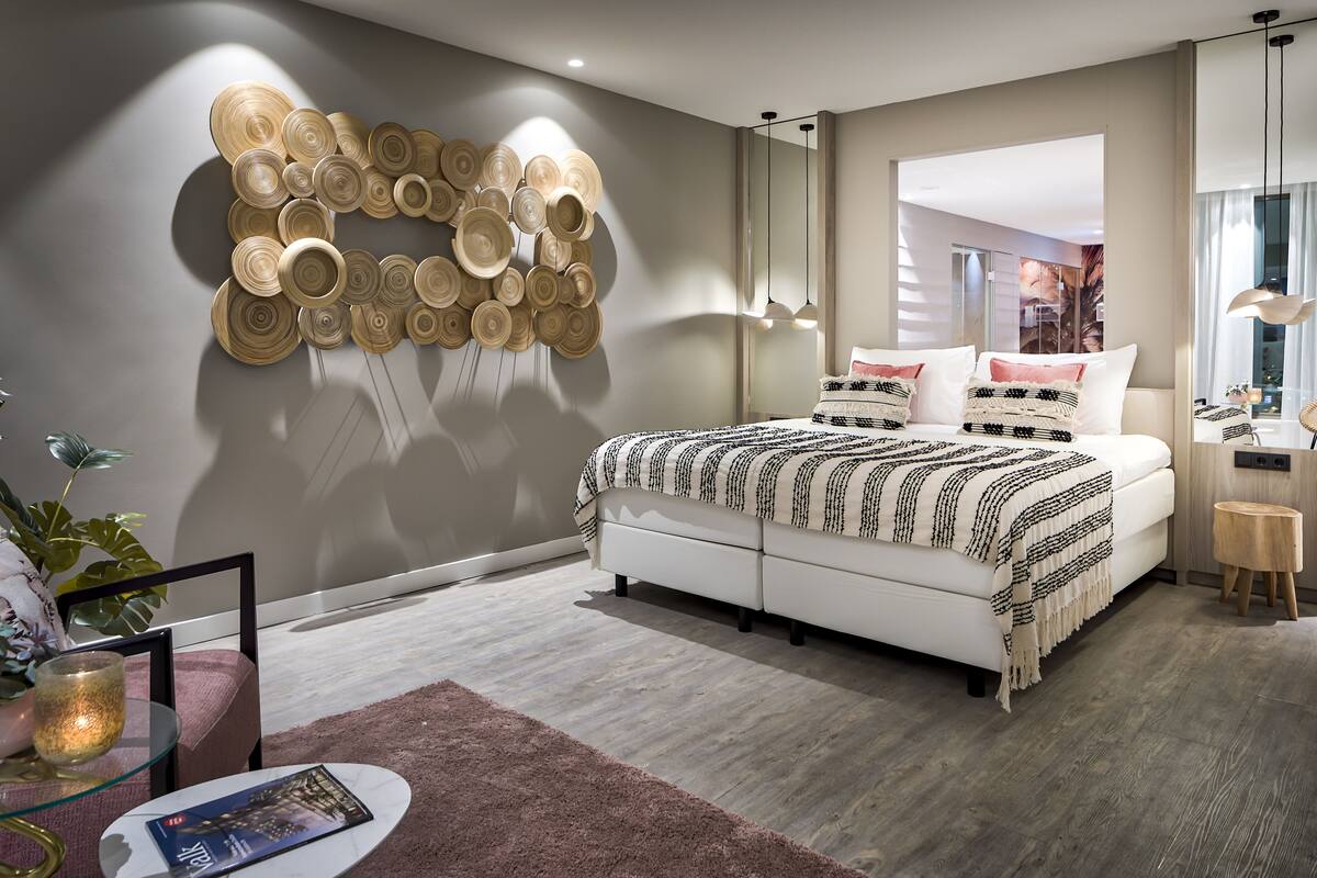 A modern bedroom with a large bed, a wall-mounted circular wall art, a small round table with a candle, and a view of the outside through a window.