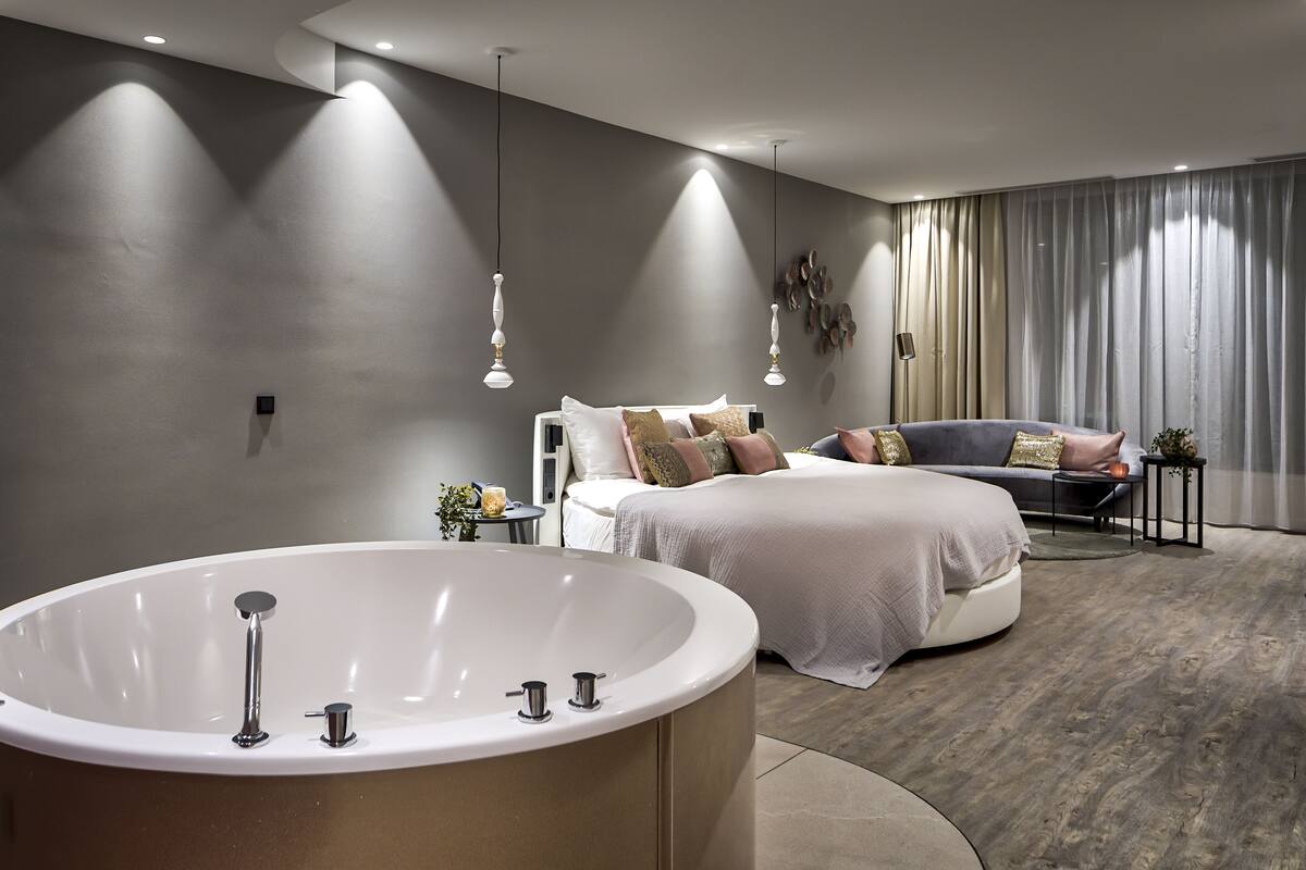 A modern bedroom with a large bathtub, a bed with multiple pillows, and a sofa.