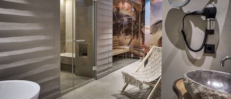 Un baño moderno con un lavabo grande blanco, cabina de ducha de vidrio y una zona de estar con una silla de macramé.