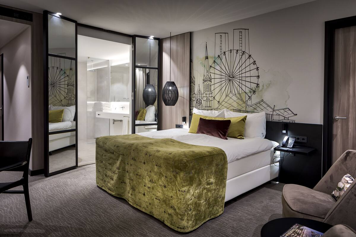 A modern hotel room with a large bed, a desk, and a chair. There is a large mirror and a wall decoration of a Ferris wheel.
