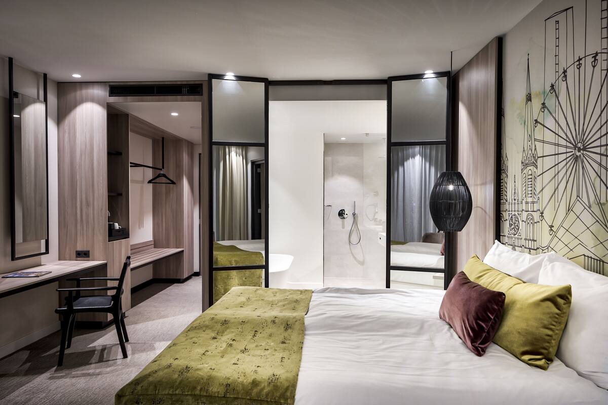 A modern hotel room with a bed, desk, chair, and a large mirror.