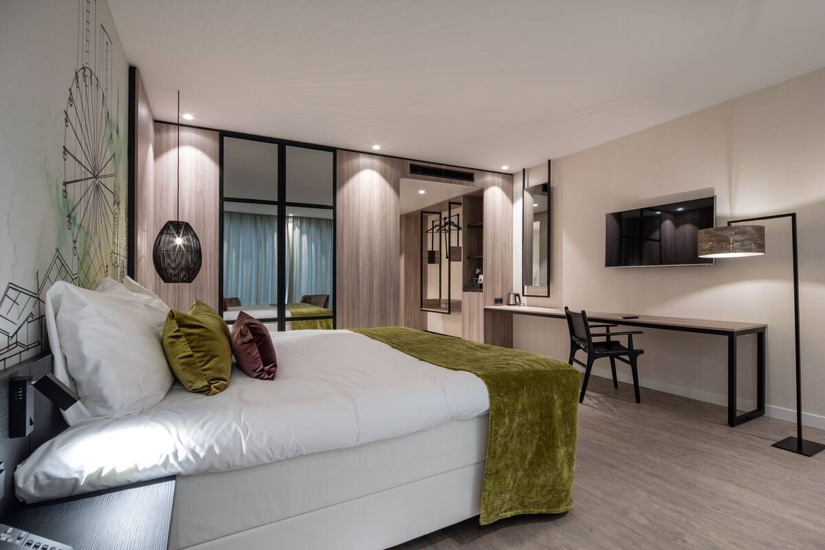 A modern hotel room with a large bed, a desk, a TV, and a mirror.