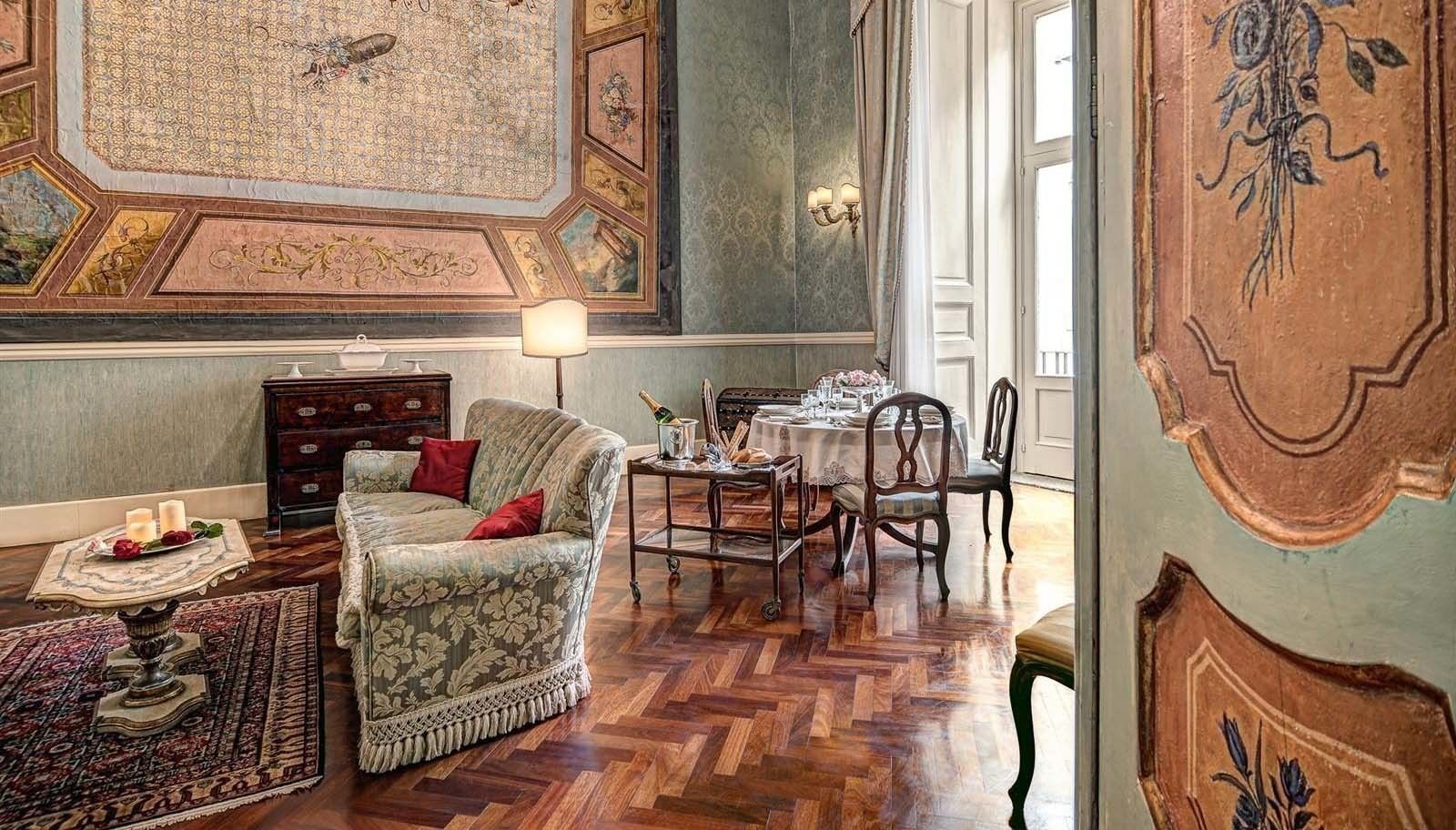 A room with a patterned carpet, a sofa with red cushions, a dining table set with chairs, and a chandelier.