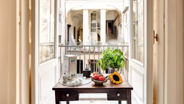 Une vue à travers une fenêtre donnant sur un couloir où l’on aperçoit une table sur laquelle sont disposés une boîte, un panier de tomates, un tournesol et une plante en pot.