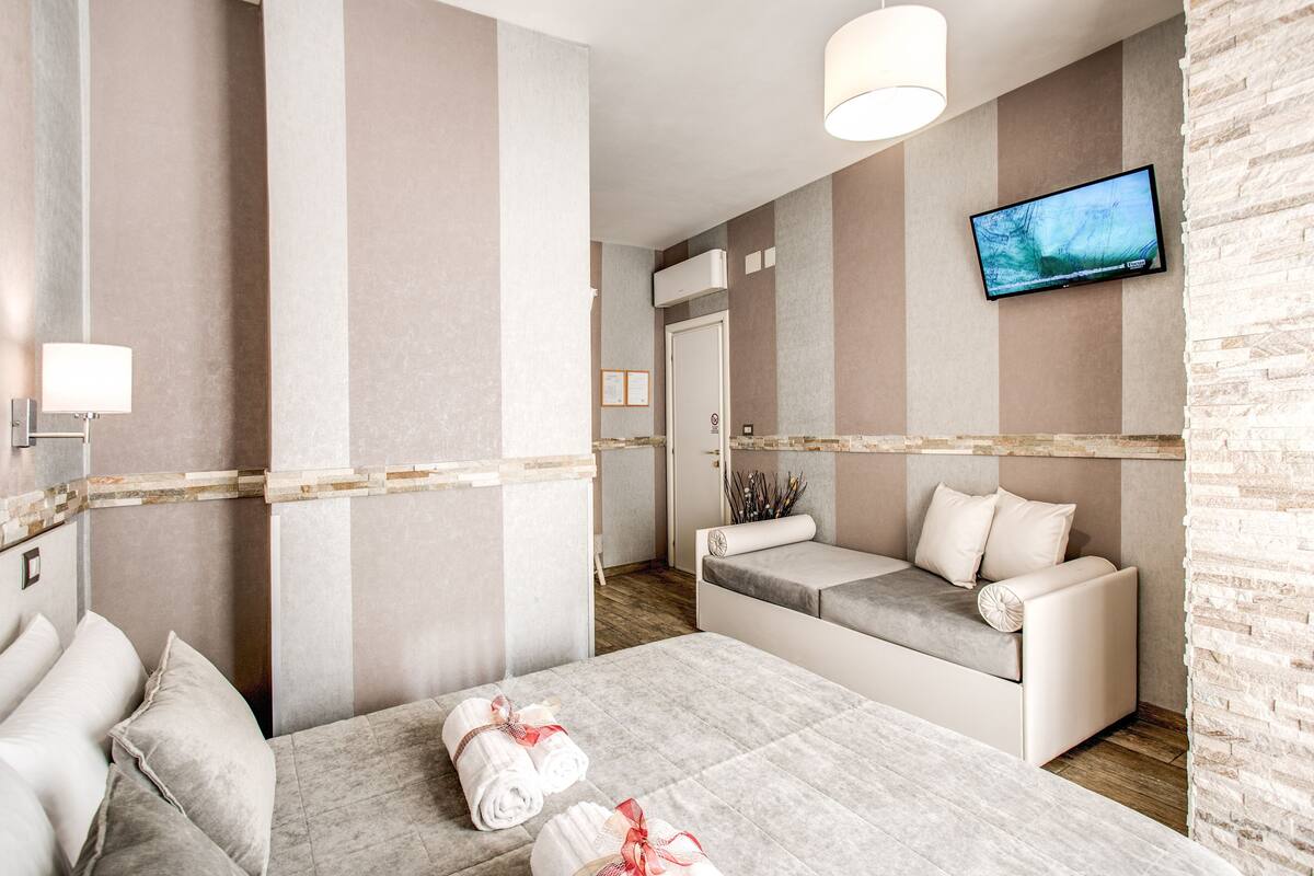 A hotel room with a large bed, a sofa, a TV mounted on the wall, and a wooden floor.