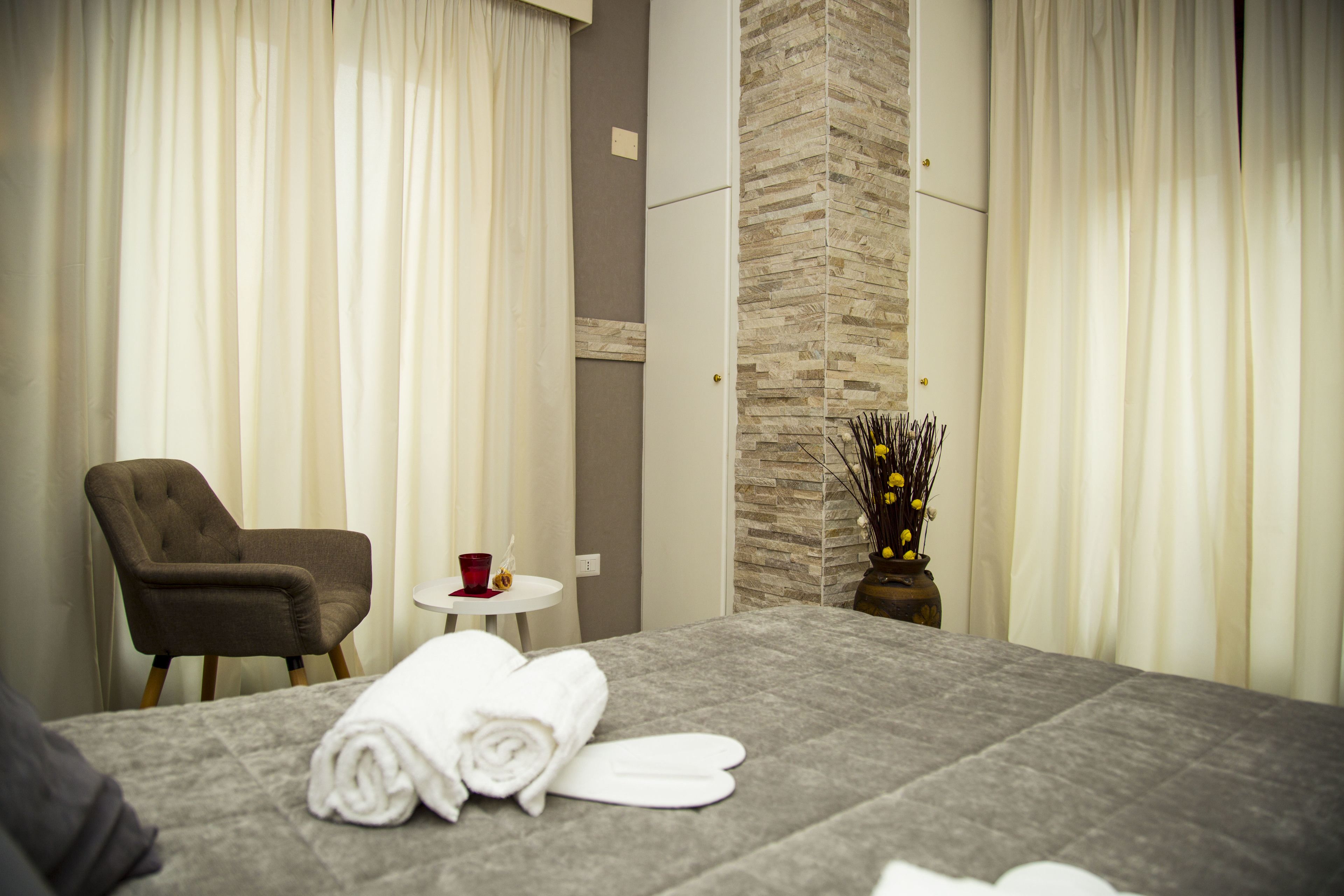 A hotel room with a bed, a chair, a small table with a red cup, and a vase with flowers.