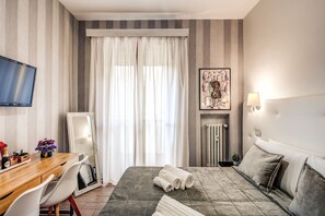 Habitación de hotel con cama, escritorio con silla, televisión, radiador y ventana con cortinas.