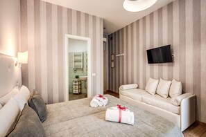 Habitación de hotel con papel tapiz a rayas, una cama, un sofá, un televisor y un baño visible a través de una puerta abierta.