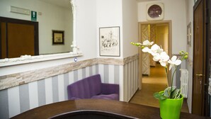 A reception area with a wooden reception desk, a green vase with white flowers, a purple chair, a mirror, and a framed picture on the wall. - Santamaria Inn (Rome)