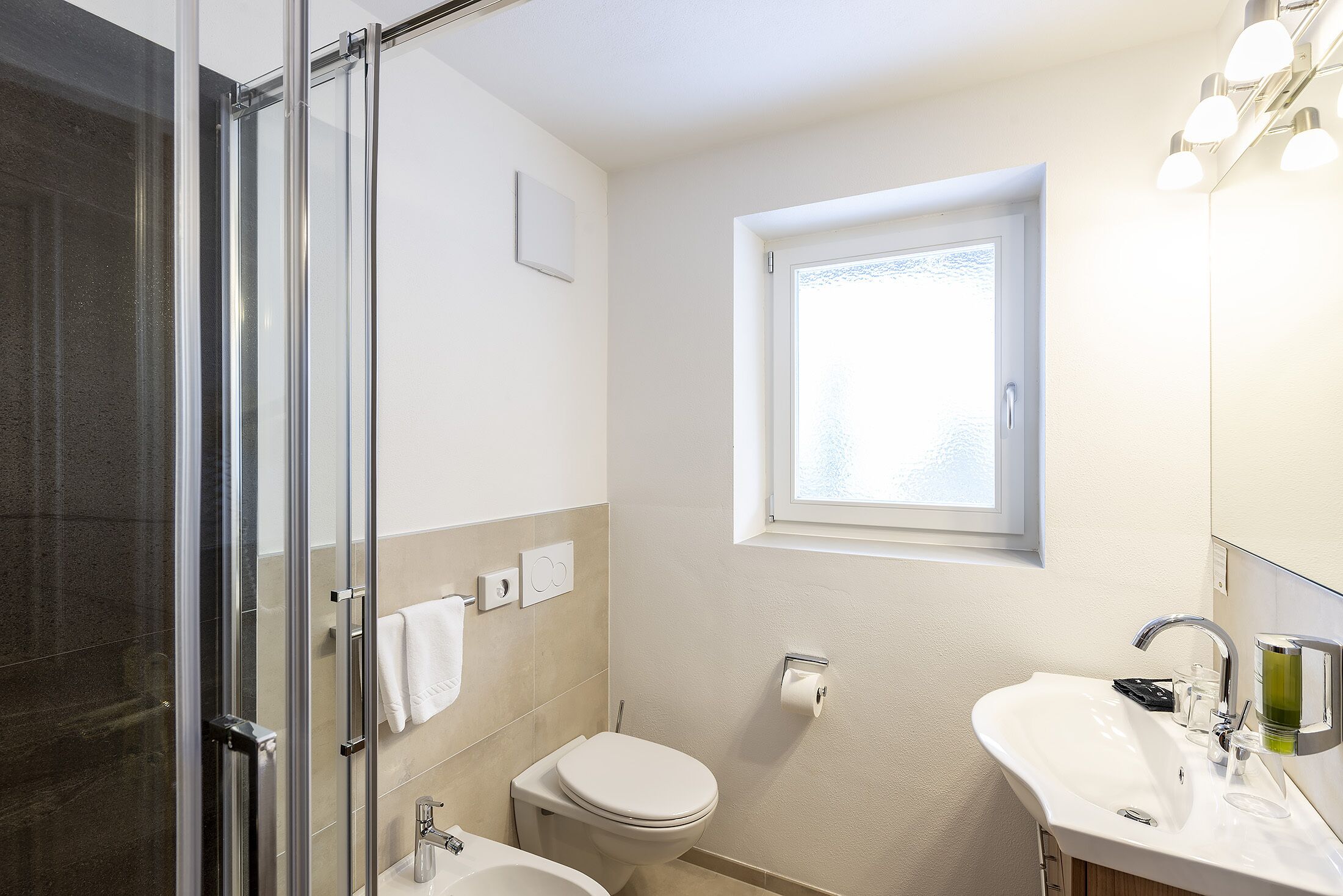 A modern bathroom with a glass shower enclosure, a toilet, a sink, and a window providing natural light.