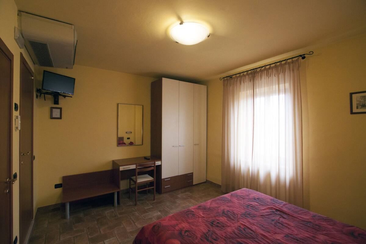 A bedroom with a bed, a desk, a wardrobe, a mirror, and a window with curtains.