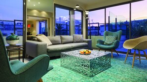 A modern living room with a grey sofa, teal armchairs, and a coffee table with fruit. Large windows offer a view of the cityscape. - Nehô Suites Cannes Croisette (Cannes)