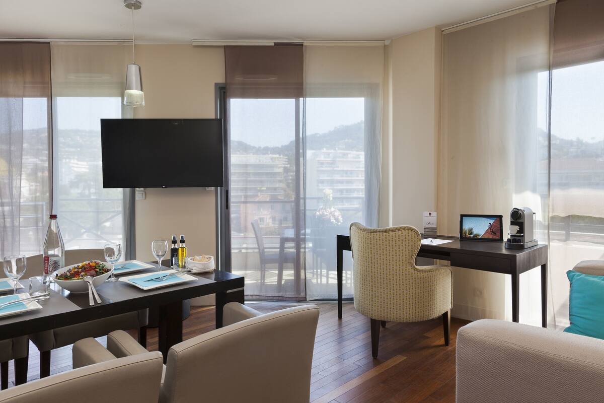 A modern hotel room with a dining area, a flat-screen TV, a desk with a computer, and a balcony with a city view.