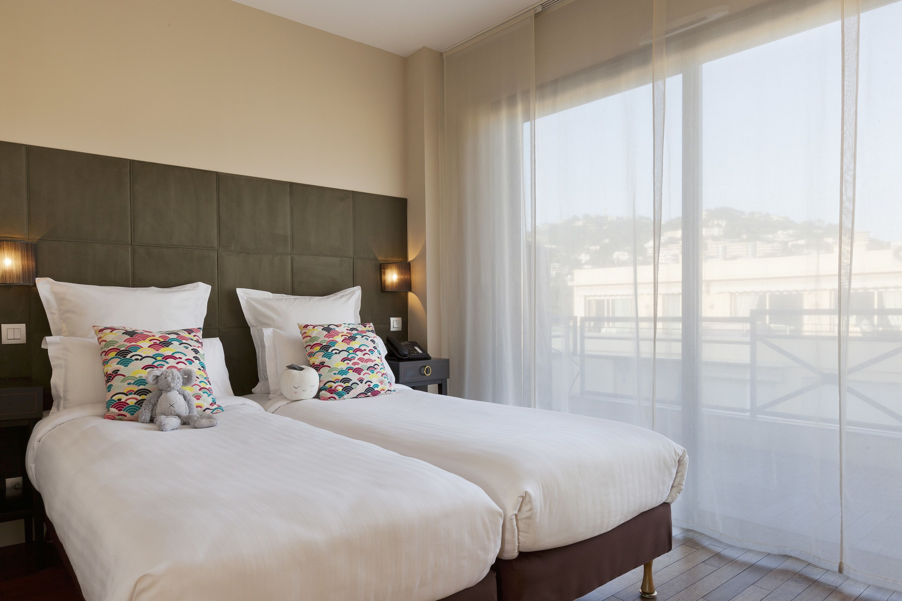 A hotel room with two beds, a large window with sheer curtains, and a view of a cityscape.