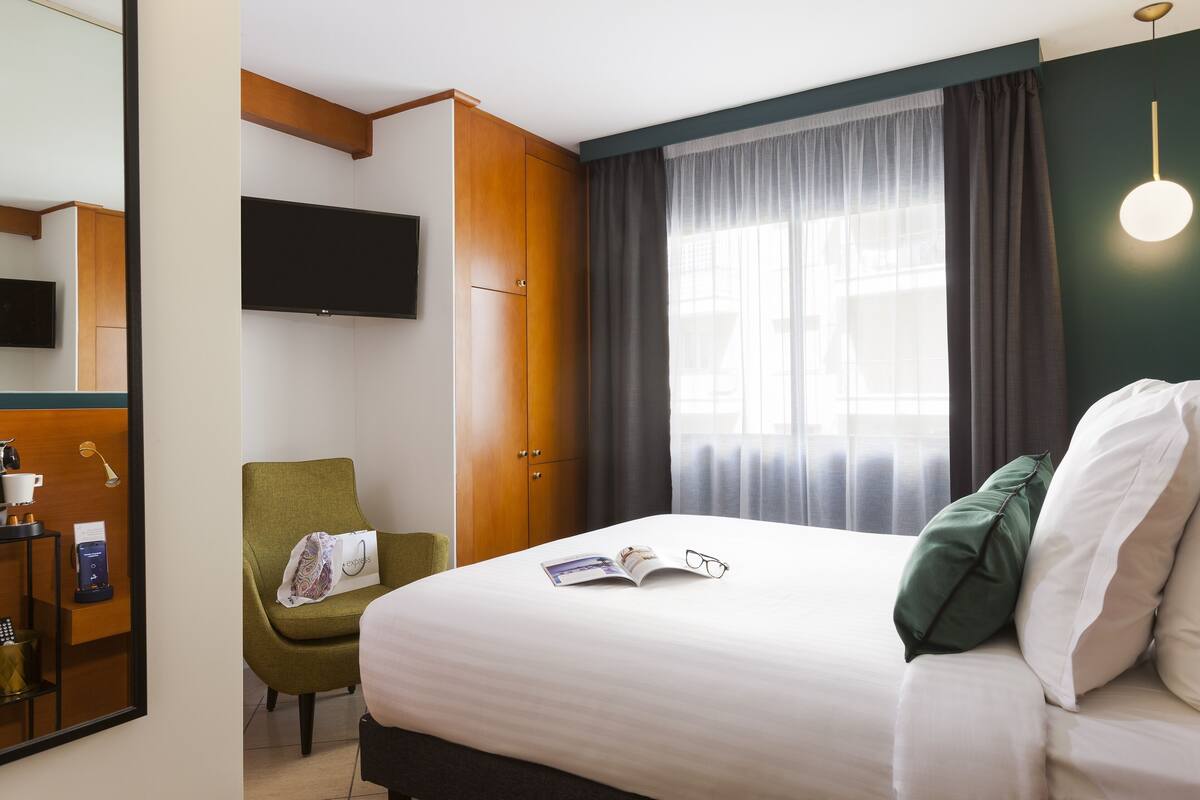 A hotel room with a bed, a green armchair, a television, and a window with curtains.