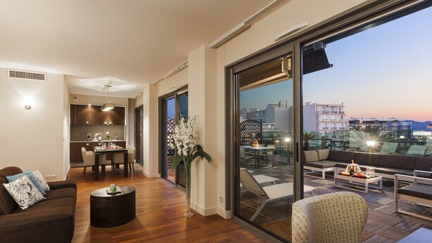 A modern living area with a sofa, coffee table, and dining table. There is a large window offering a view of the cityscape. - Nehô Suites Cannes Croisette (Cannes)