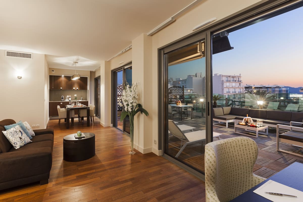 A modern living area with a sofa, coffee table, and dining table. There is a large window offering a view of the cityscape.