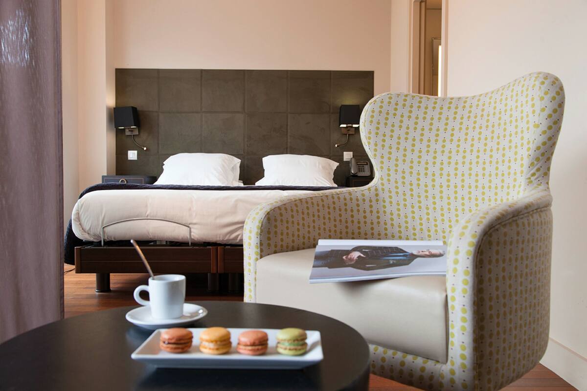 A hotel room with a bed, a chair, a table with a coffee cup and macarons, and a magazine.