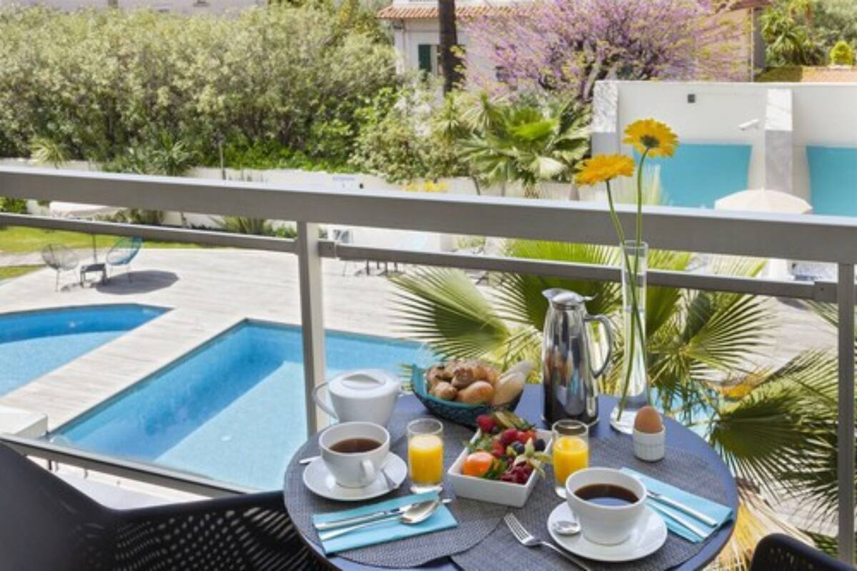A balcony with a breakfast setup including coffee, juice, and fruit, overlooking a swimming pool and lush greenery.