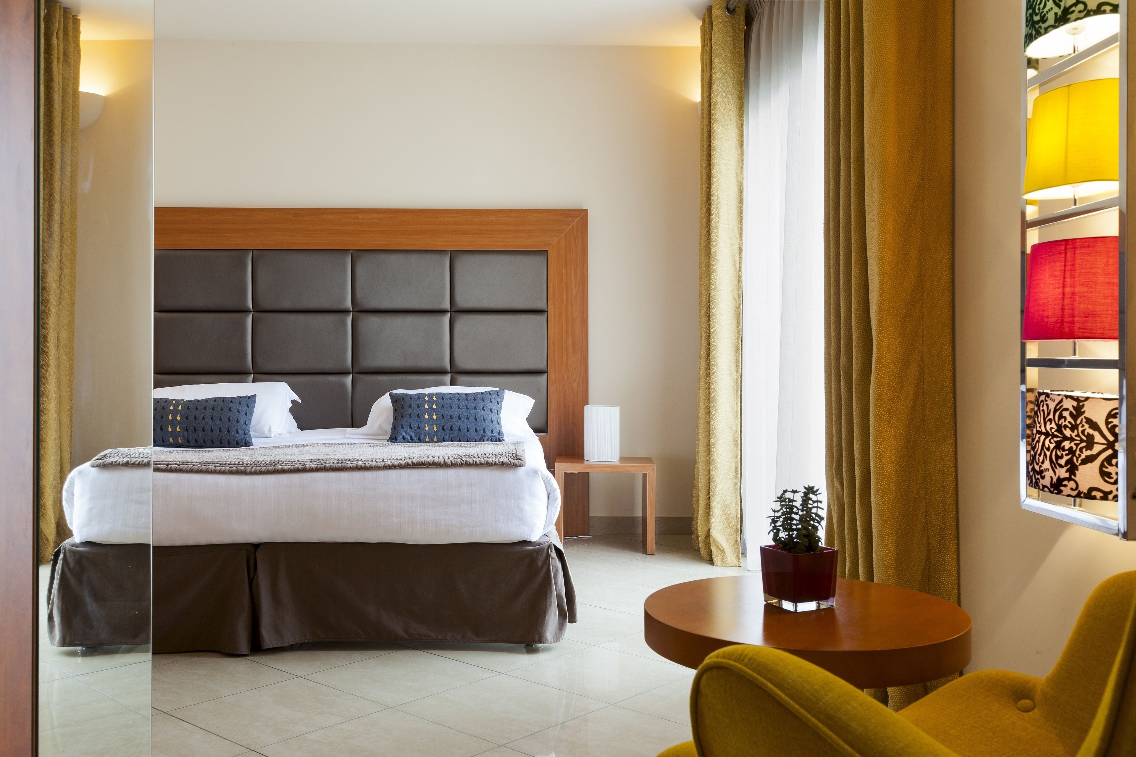 A modern hotel room with a large bed, a bedside table, a small table with a plant, and a window with curtains.