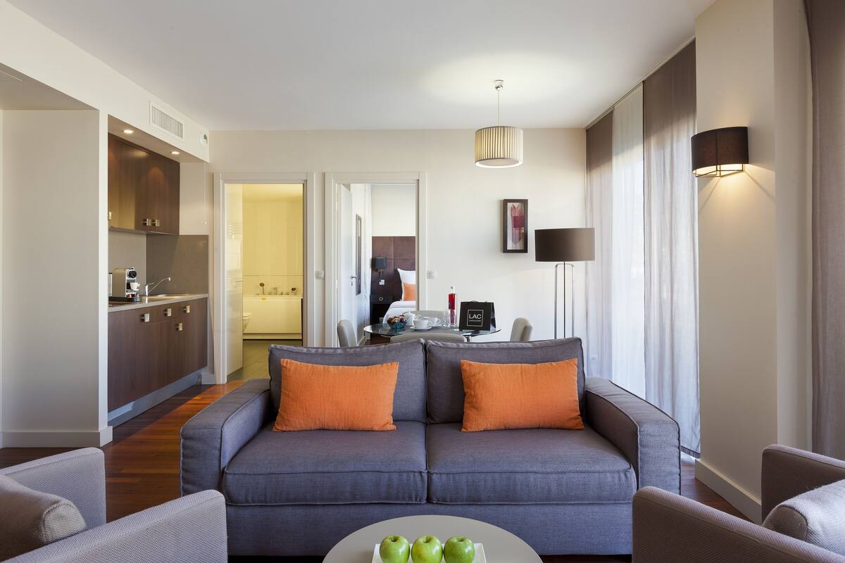 A modern living room with a grey sofa, orange cushions, and a coffee table with apples. There is a kitchen area with wooden cabinets and a dining table in the background.