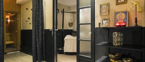 A room with a wooden floor, a black curtain, a bed, a shelf with religious items, and a wall with framed pictures.