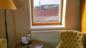 A room with two patterned armchairs, a small round table with a tray of items, a lamp, and a window with a view of a building and a red roof. - Hotel De Valk (Westkapelle)