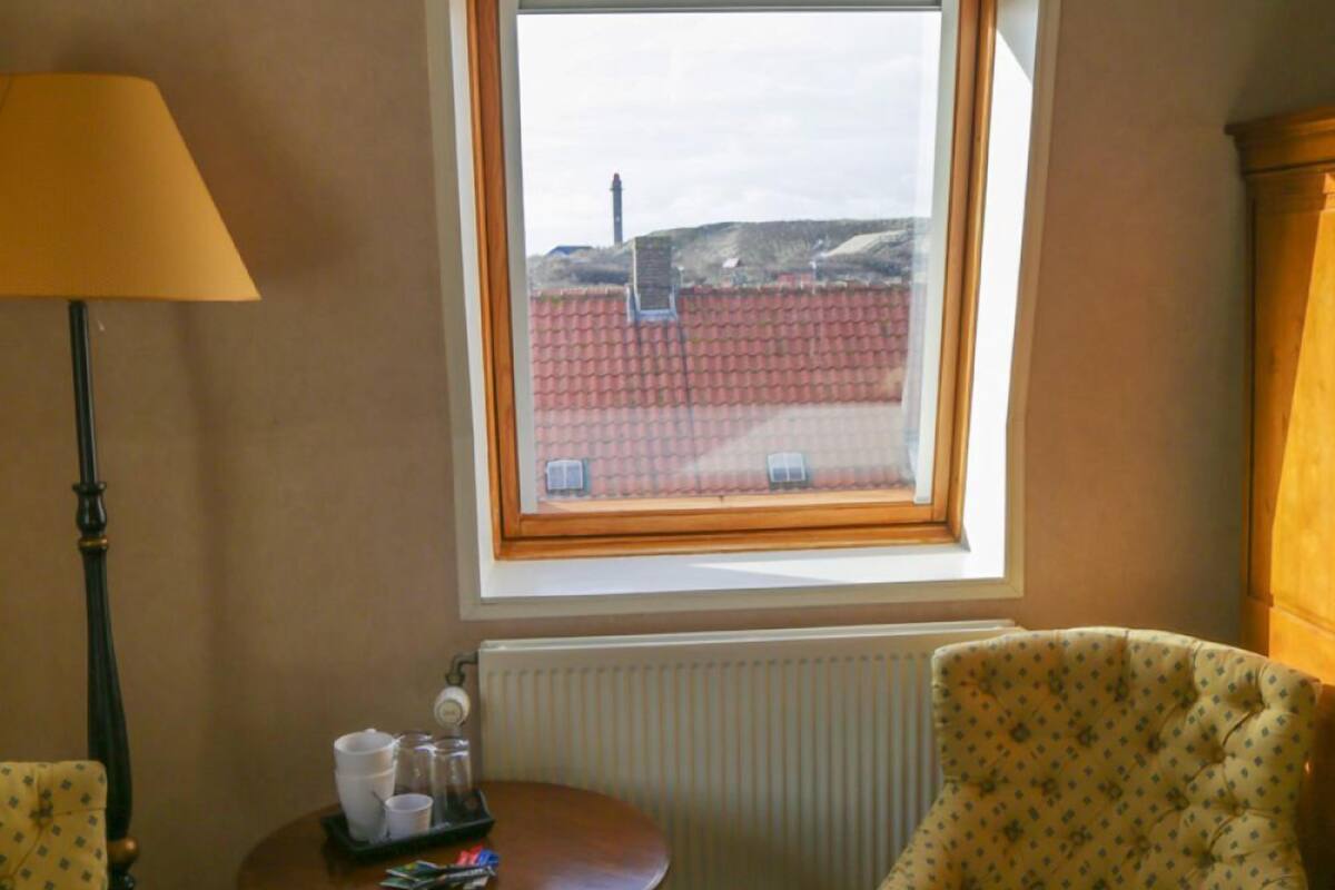 Een kamer met twee gestoffeerde fauteuils, een kleine ronde tafel met een dienblad en spullen, een lamp en een raam met uitzicht op een gebouw en een rood dak.