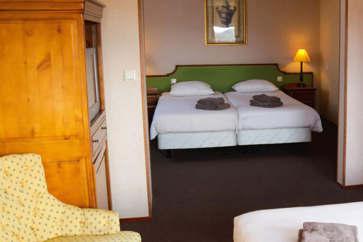 Een hotelkamer met een tweepersoonsbed, een houten garderobe, een groen hoofdeinde, een ingelijste foto aan de muur en een bedlampje.