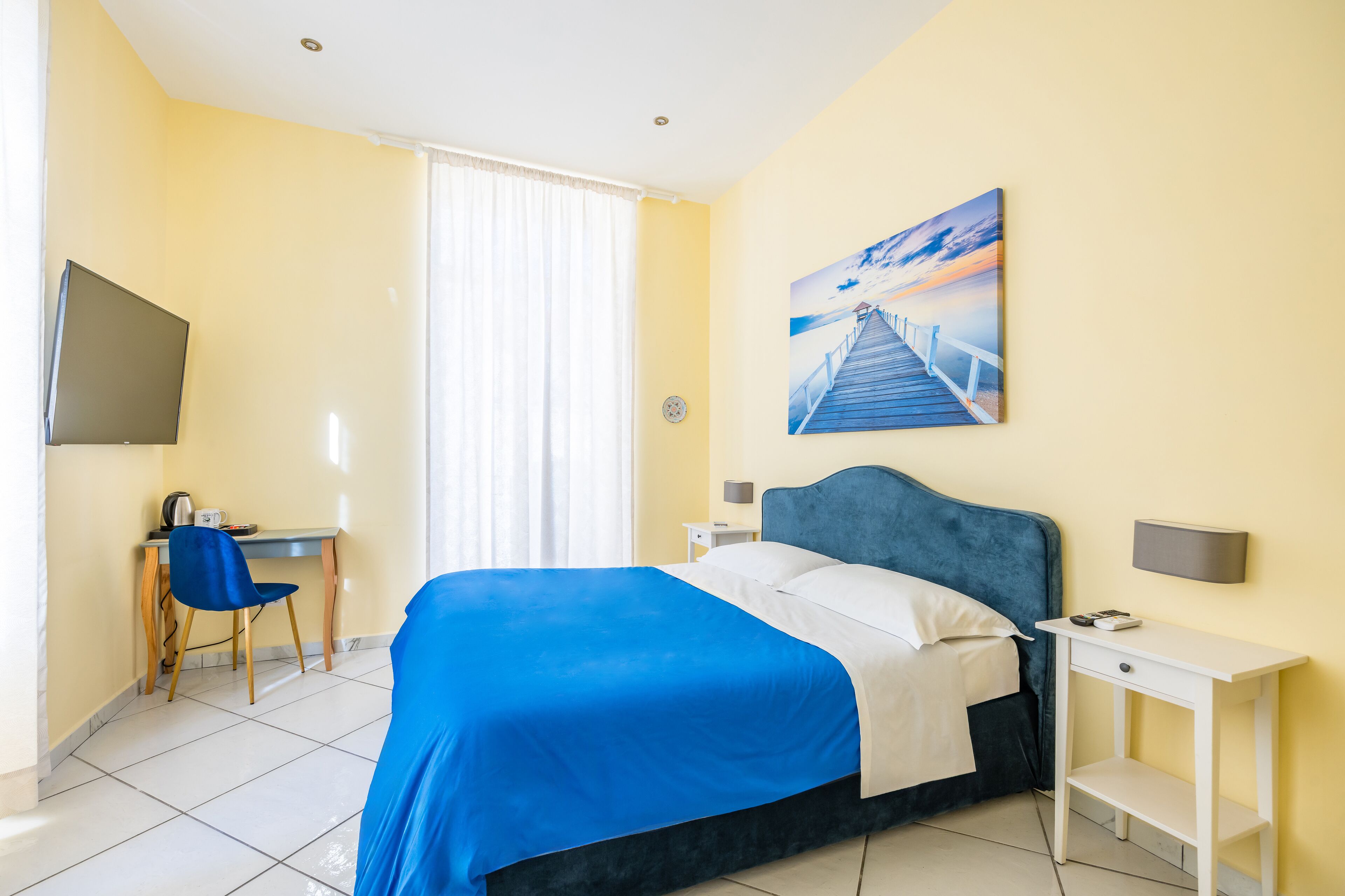 A hotel room with a bed, a desk with a chair, a TV, and a painting on the wall.
