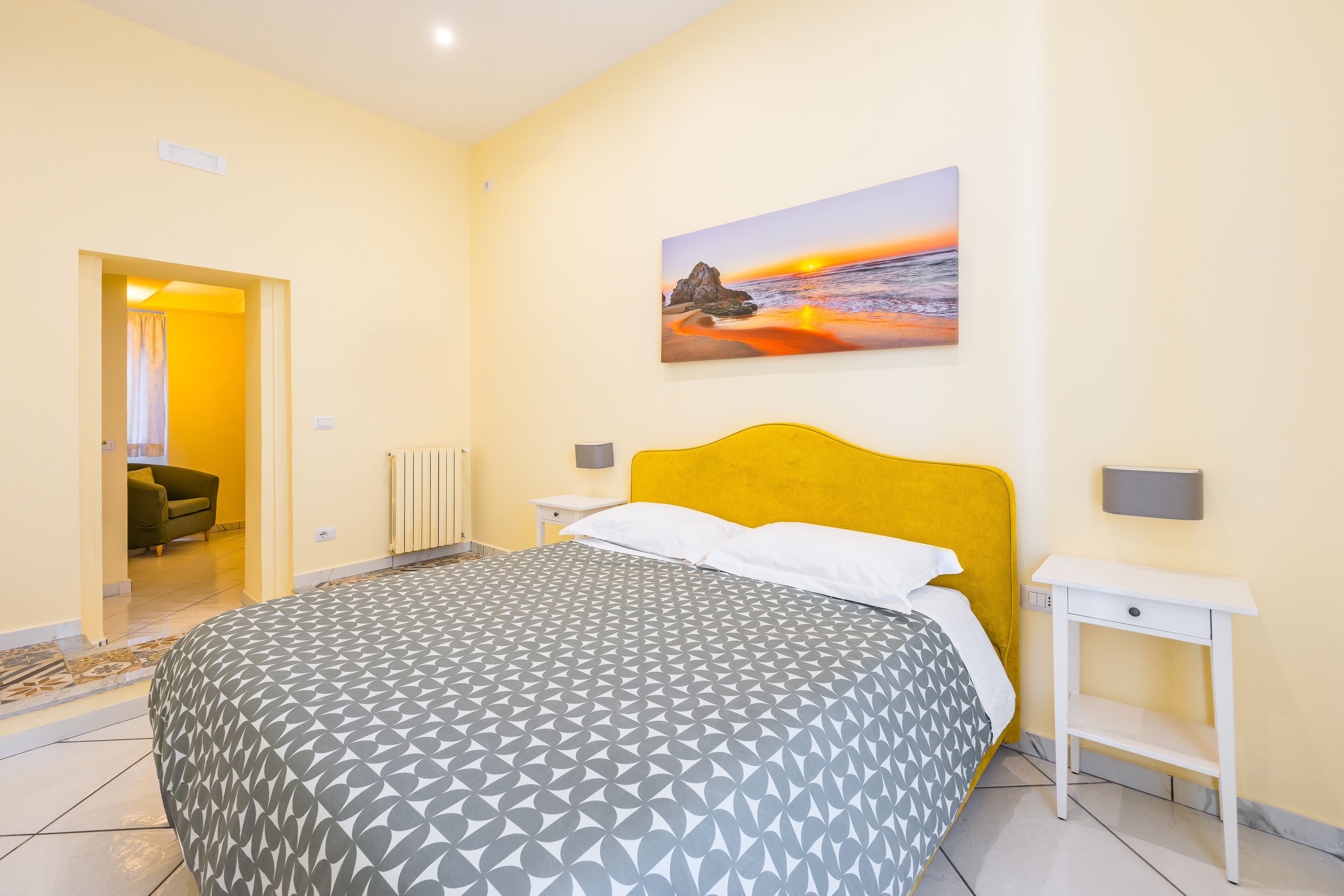A bedroom with a bed, a nightstand, a painting of a sunset, and a bathroom visible through an open door.