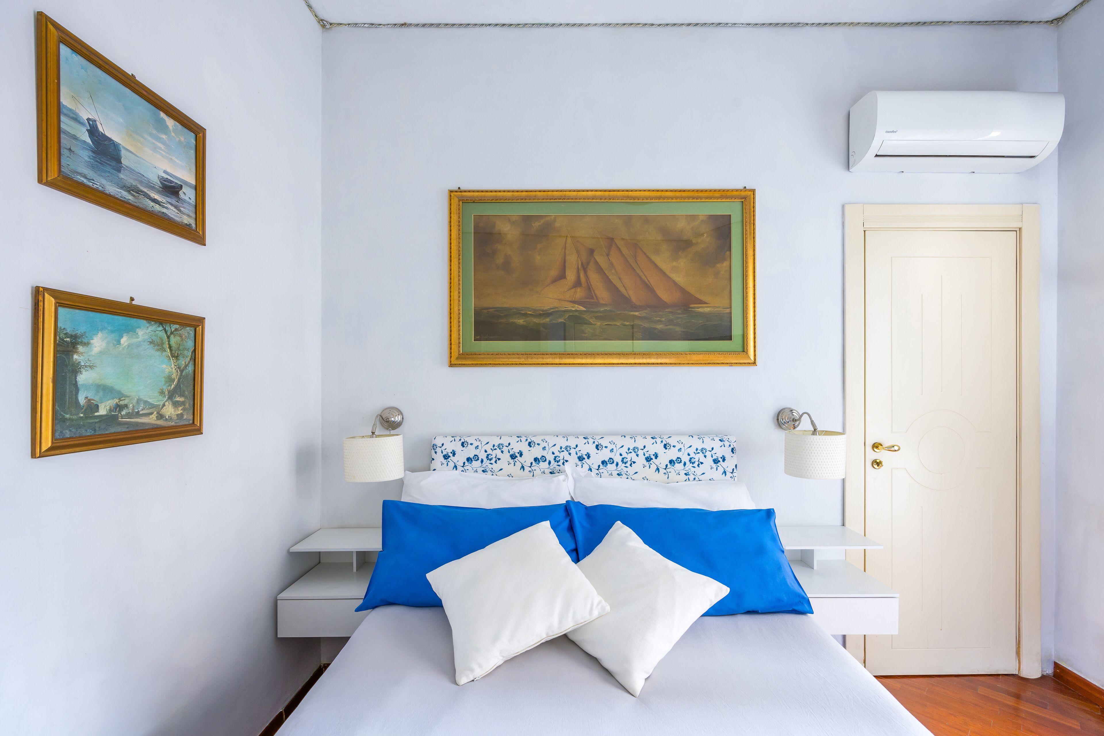 A bedroom with a bed, two pillows, a nightstand, a lamp, a wall-mounted air conditioner, and two framed paintings.