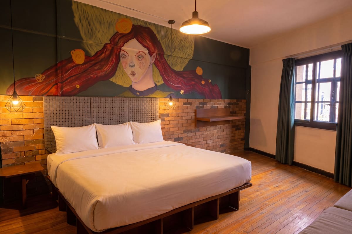 Habitación de hotel con una cama grande, un muro decorado con ladrillos, un mural y una ventana con cortinas.