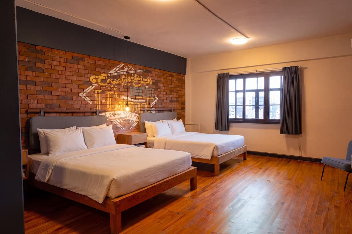 Una habitación con dos camas, una pared de ladrillo con grafitis, una ventana con cortinas y un piso de madera.