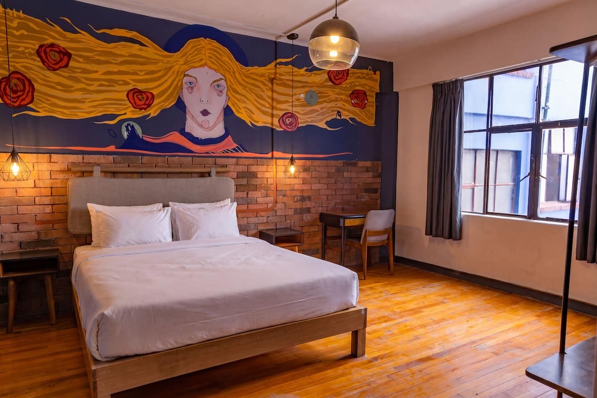 Un dormitorio con una cama, pared de ladrillo, mural, ventana y una mesa.
