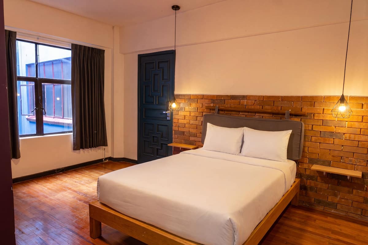 Un dormitorio con cama, suelo de madera, una ventana con cortinas, una puerta azul y una pared de ladrillo.
