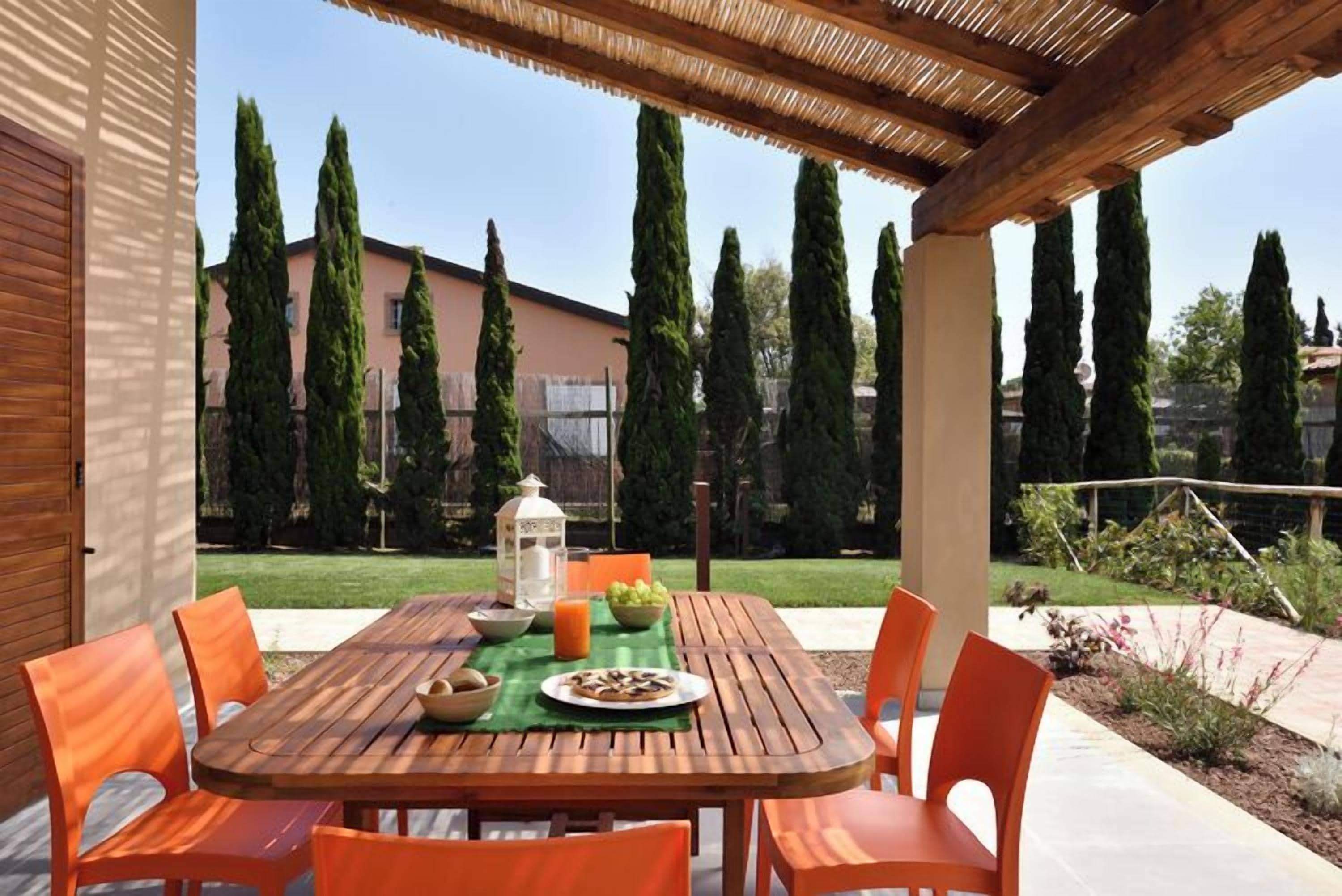 An outdoor patio with a wooden table and orange chairs, a lantern, and a bowl of fruit.