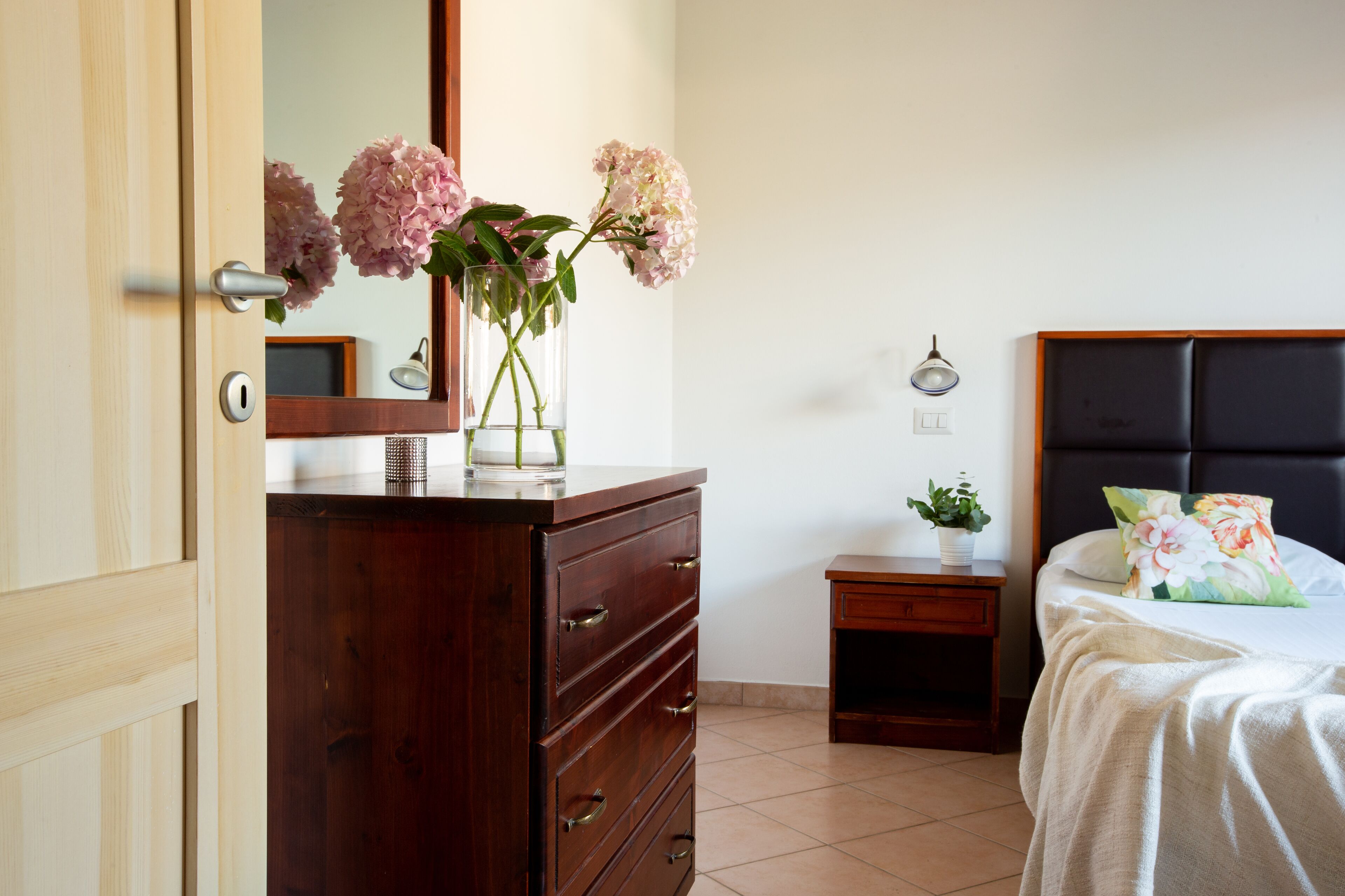 A bedroom with a wooden dresser, a bedside table with a lamp, a bed with a floral pillow, and a mirror on the wall.