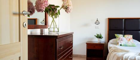 A bedroom with a wooden dresser, a bedside table with a lamp, a bed with a floral pillow, and a mirror on the wall.