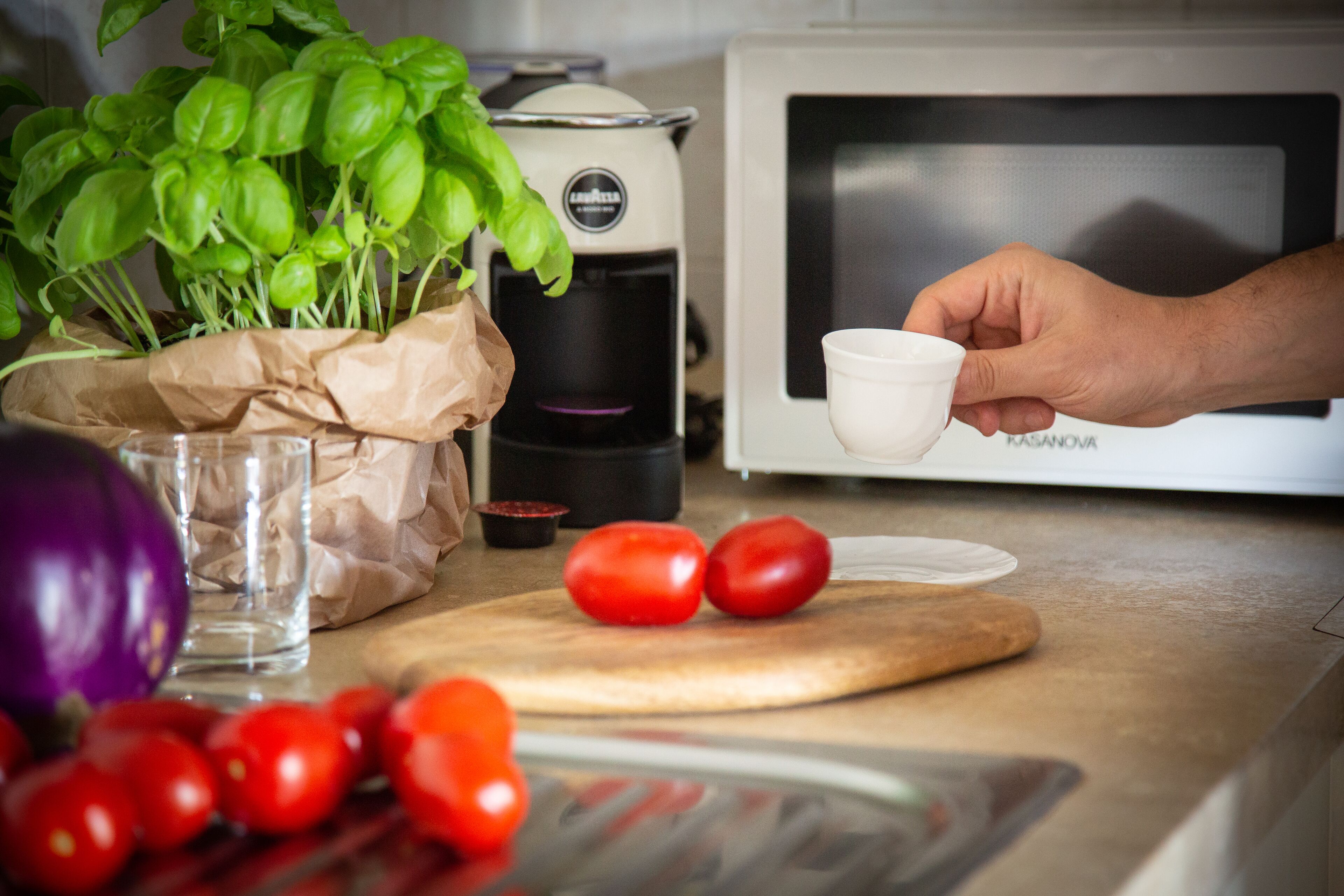 A kitchen counter with a coffee maker, a microwave, a cutting board with tomatoes, and a hand holding a cup.