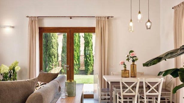 A modern living room with a sofa, a wooden table, and a dining area with white chairs. There are hanging lights and a view of greenery outside. - Borgo Verde (Rosignano Marittimo)